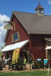 A red barn-style building with a standing seam metal roof, featuring a cupola and weather vane, surrounded by outdoor furniture and "New Showroom" banners.
