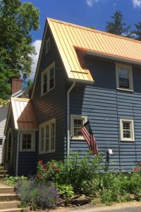 A blue house with a newly installed copper-colored standing seam metal roof, complemented by a small garden and an American flag in the foreground.