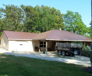 A beige home with a newly installed standing seam metal roof in a rich brown color, surrounded by green trees and a driveway with a utility trailer parked nearby.