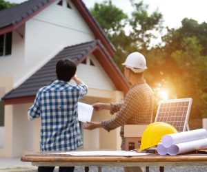 Two men discussing a roof installation in front of a home with roofing plans and tools on a table.