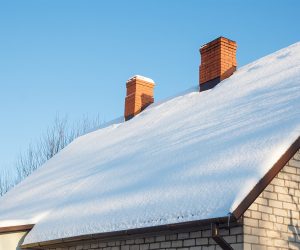 Snow-covered roof with three red brick chimneys under a clear blue sky.