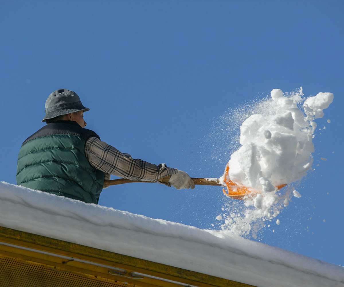 Homeowner safely shoveling heavy snow buildup off roof under clear winter sky.