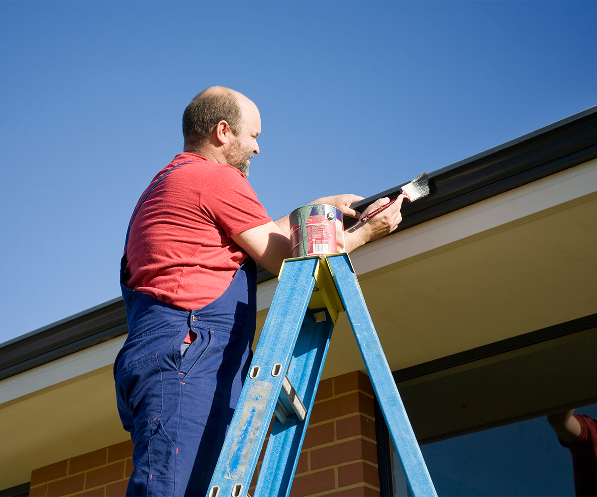 Man on ladder painting eaves and fascia as part of fall roof maintenance.