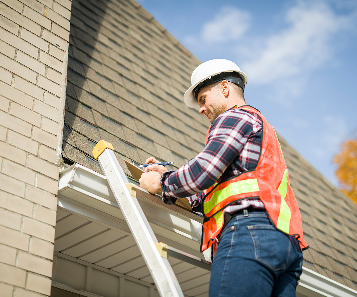 Roofing inspector on ladder checking gutters and shingles during professional fall roof inspection.